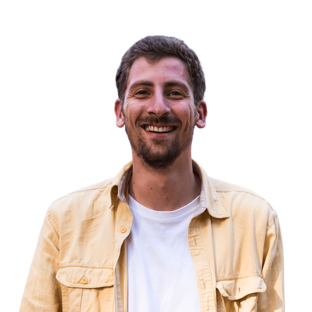 Een man met kort bruin haar en een baard glimlacht, draagt een lichtgeel overhemd over een wit t-shirt en staat tegen een effen witte achtergrond.