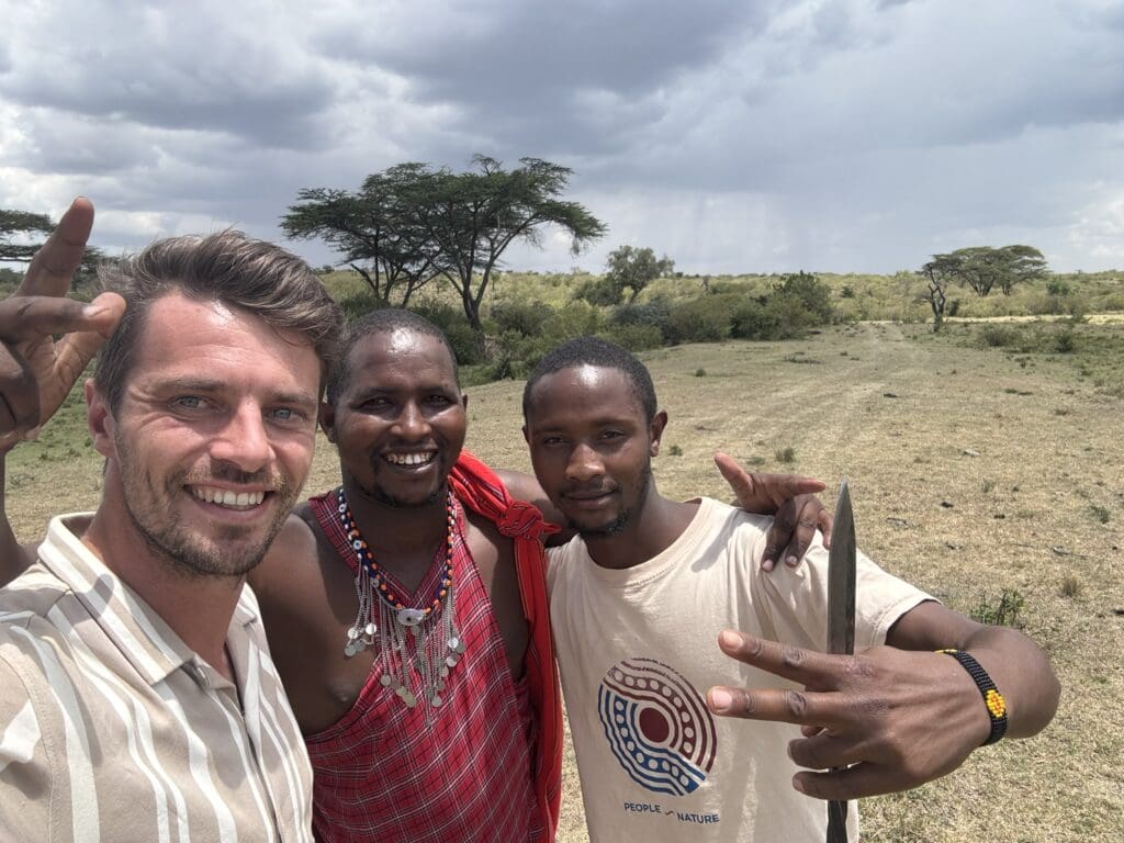 Drie mannen lachen en poseren voor een selfie in een open graslandschap met bomen. Eén man draagt traditionele Maasai kleding, terwijl de anderen casual shirts dragen. Twee van hen maken vredestekens met hun handen onder een bewolkte hemel.