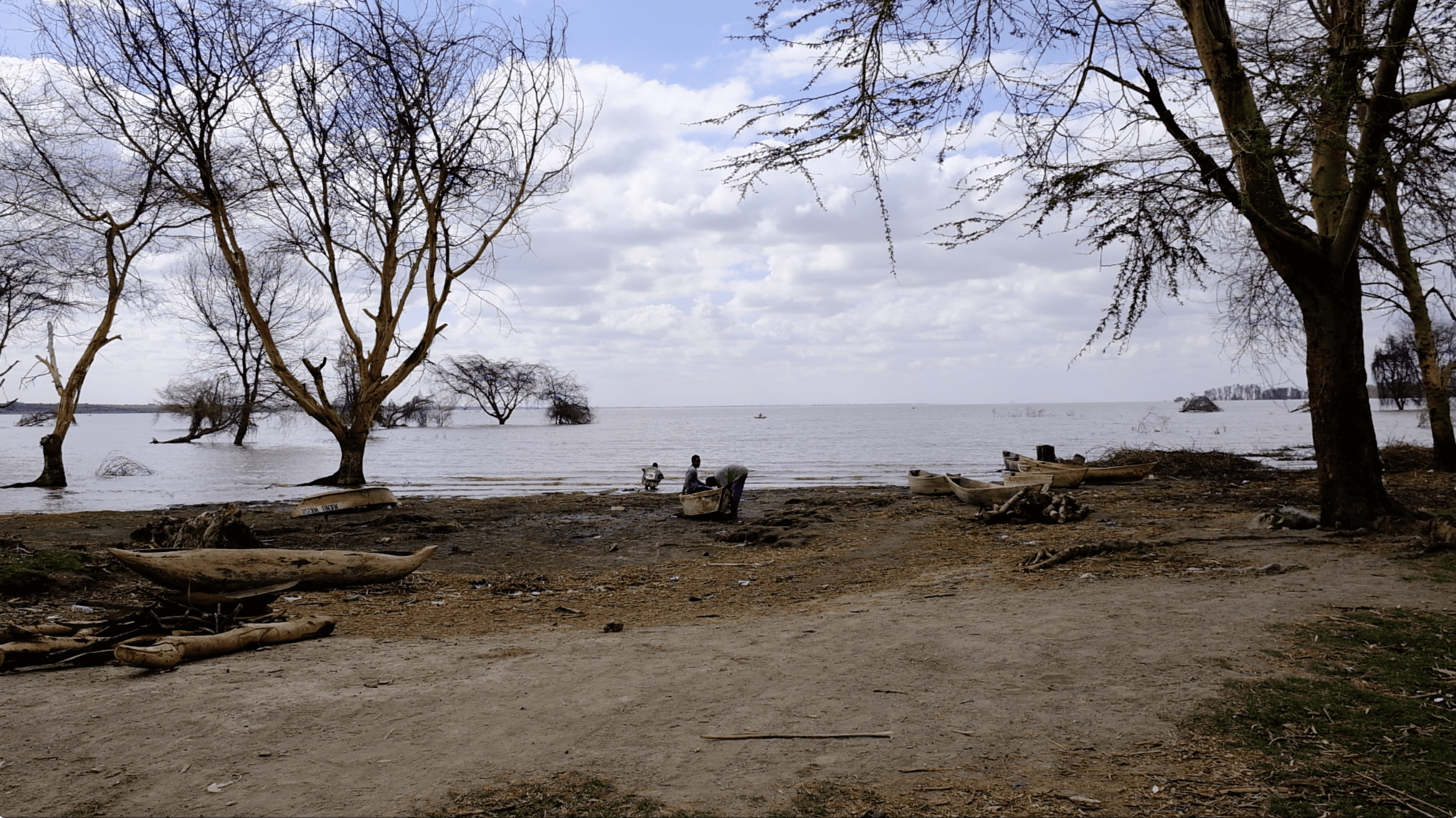 Een scène aan de oever van een meer met twee mensen bij houten boten op de zanderige, met bomen omzoomde oever onder een gedeeltelijk bewolkte hemel. Enkele bomen staan in ondiep water en het meer strekt zich uit tot aan de horizon.