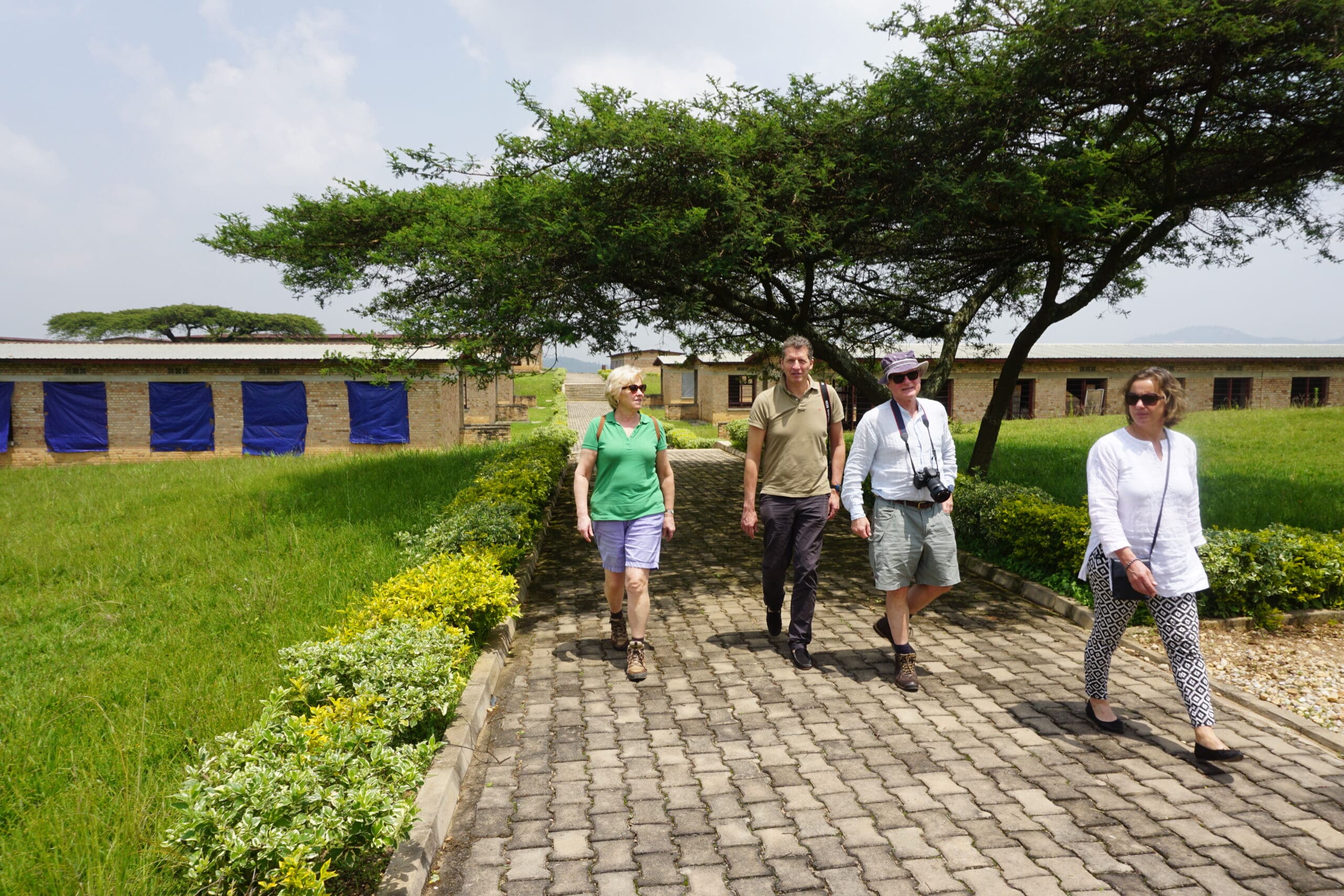 Vier mensen lopen over een geplaveid pad omzoomd door groene struiken en gras, met bomen die schaduw bieden en stenen gebouwen met blauwe deuren op de achtergrond op een zonnige dag.
