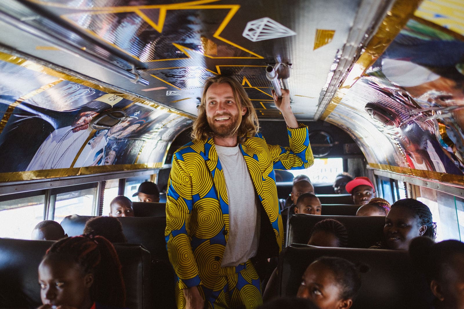Een lachende man in een fel geel en blauw pak met patroon staat in een overvolle bus met kinderen om hem heen. Het plafond van de bus is versierd met kleurrijke patronen en foto's.