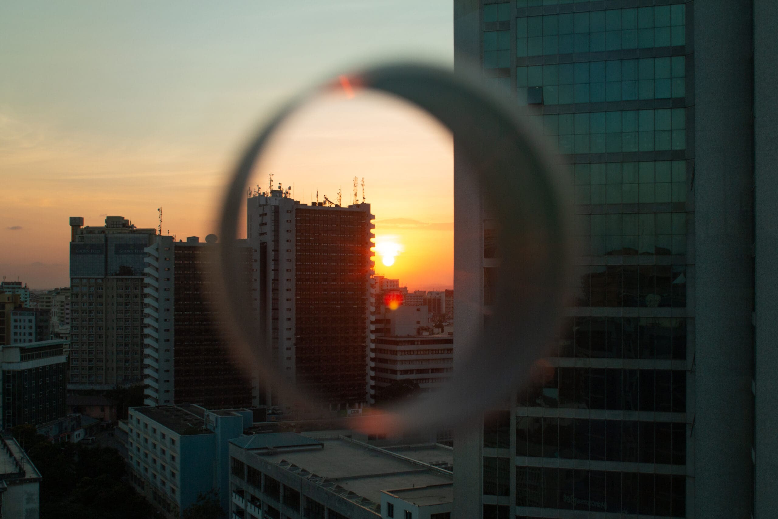 Een skyline van een stad bij zonsondergang, met hoge gebouwen in silhouetten tegen een oranje lucht. Een cirkelvormig glazen object op de voorgrond vervaagt en vervormt een deel van het uitzicht, waardoor een zacht visueel effect ontstaat.