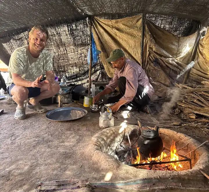 Twee mannen zitten in een tent; de ene knielt bij een klein vuurtje met een theepot, de andere glimlacht naar de camera. Kookgerei en voorraden liggen verspreid op de grond om hen heen. Zonlicht schijnt door het tentdoek naar binnen.