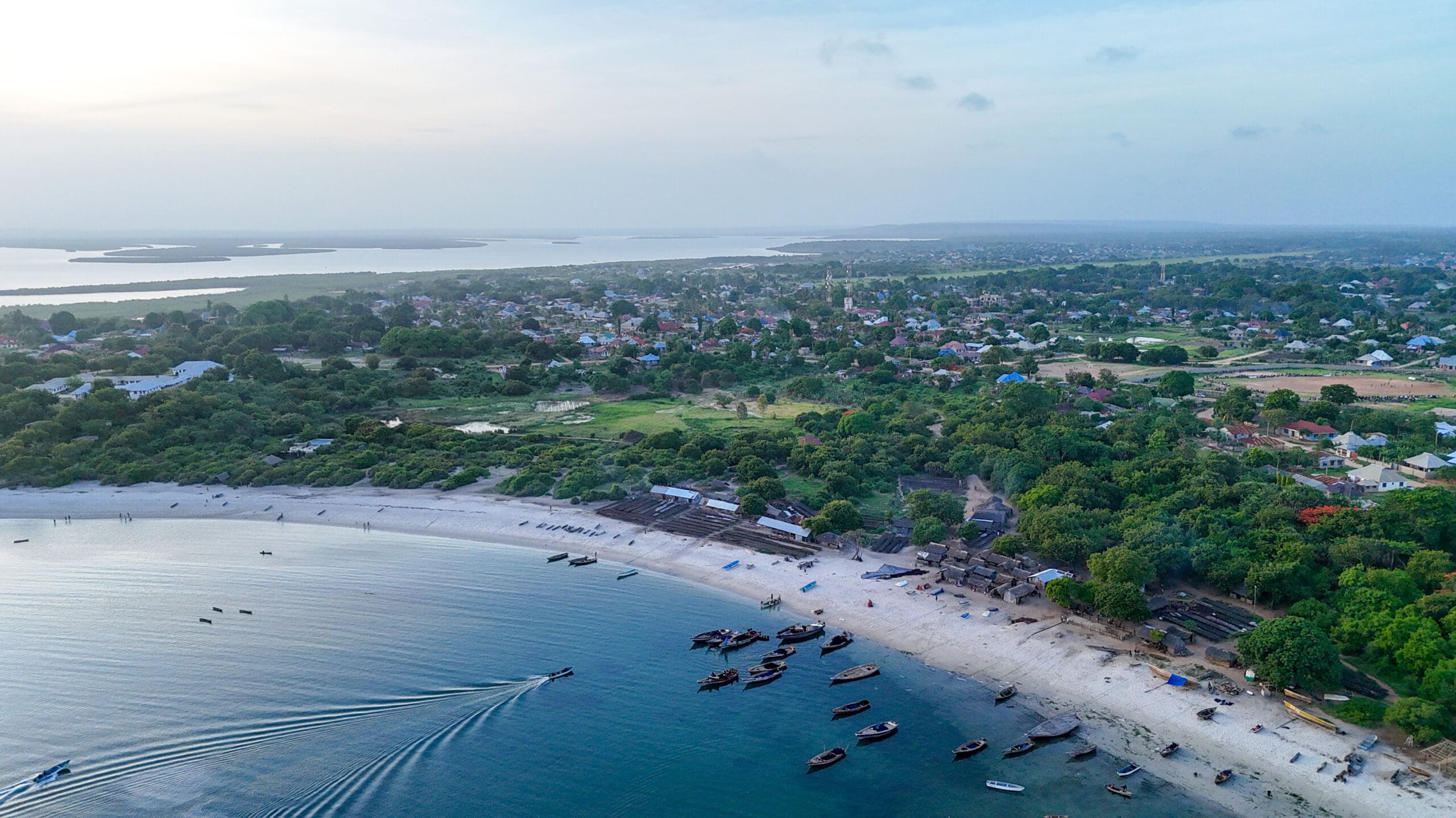 Luchtfoto van een kustplaats met weelderig groen, een zandstrand, kleine boten in het water en kalme golven, gemaakt bij zonsondergang met een zachte, blauwe lucht erboven.