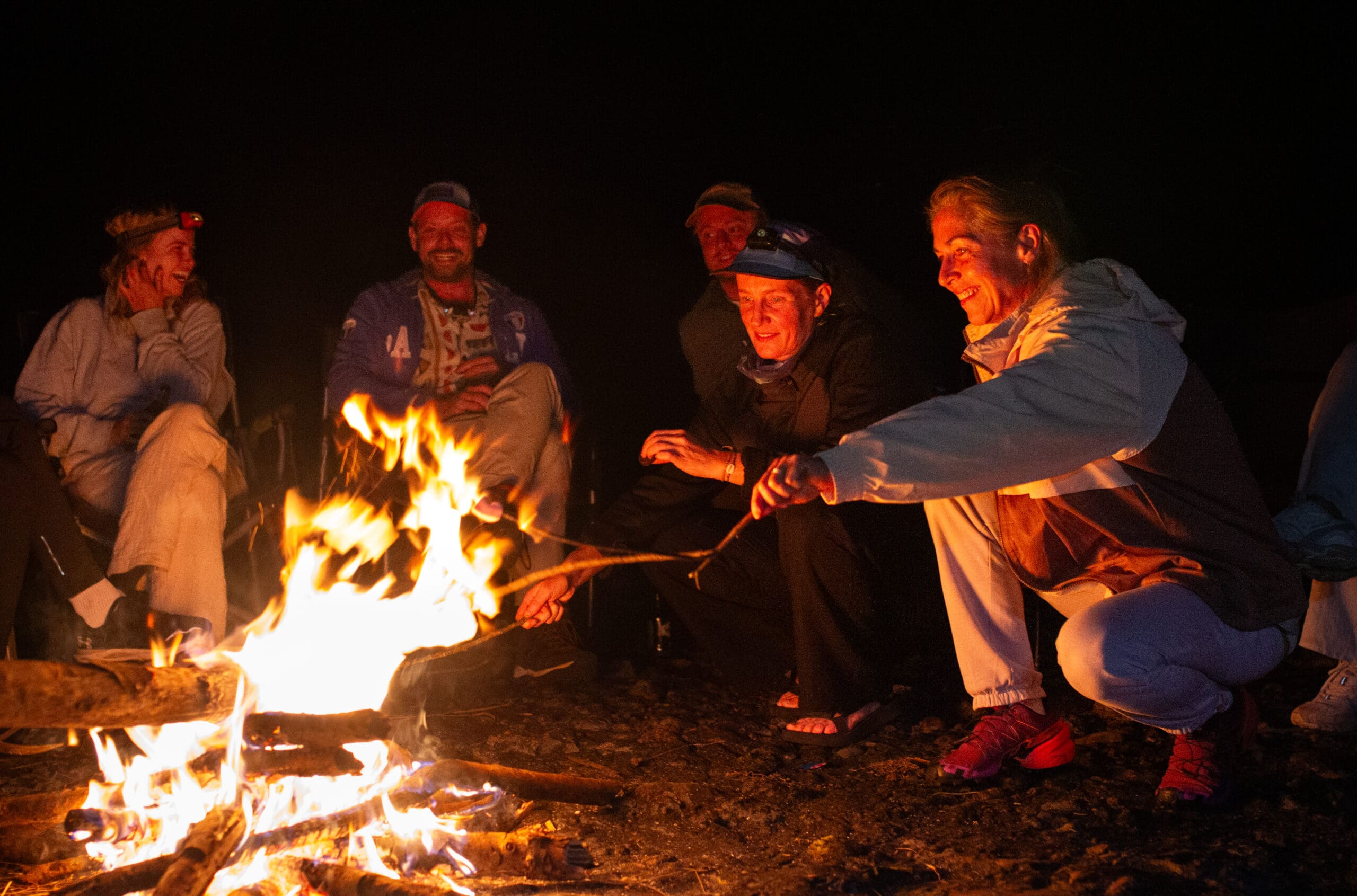 Een groep mensen zit en knielt 's nachts rond een kampvuur, glimlacht en roostert marshmallows boven de vlammen. Het warme vuur verlicht hun gezichten tegen de donkere achtergrond.