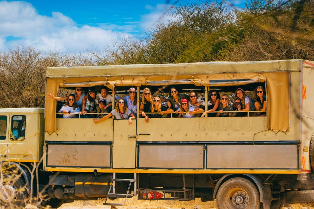 Een groep mensen glimlacht en leunt uit de ramen van een grote beige safaritruck die buiten geparkeerd staat, omringd door droge struiken onder een felblauwe hemel.