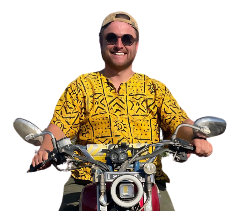 Een lachende man met zonnebril, beige pet en felgeel overhemd met patroon zit op een motorfiets, houdt het stuur vast, met een witte achtergrond.