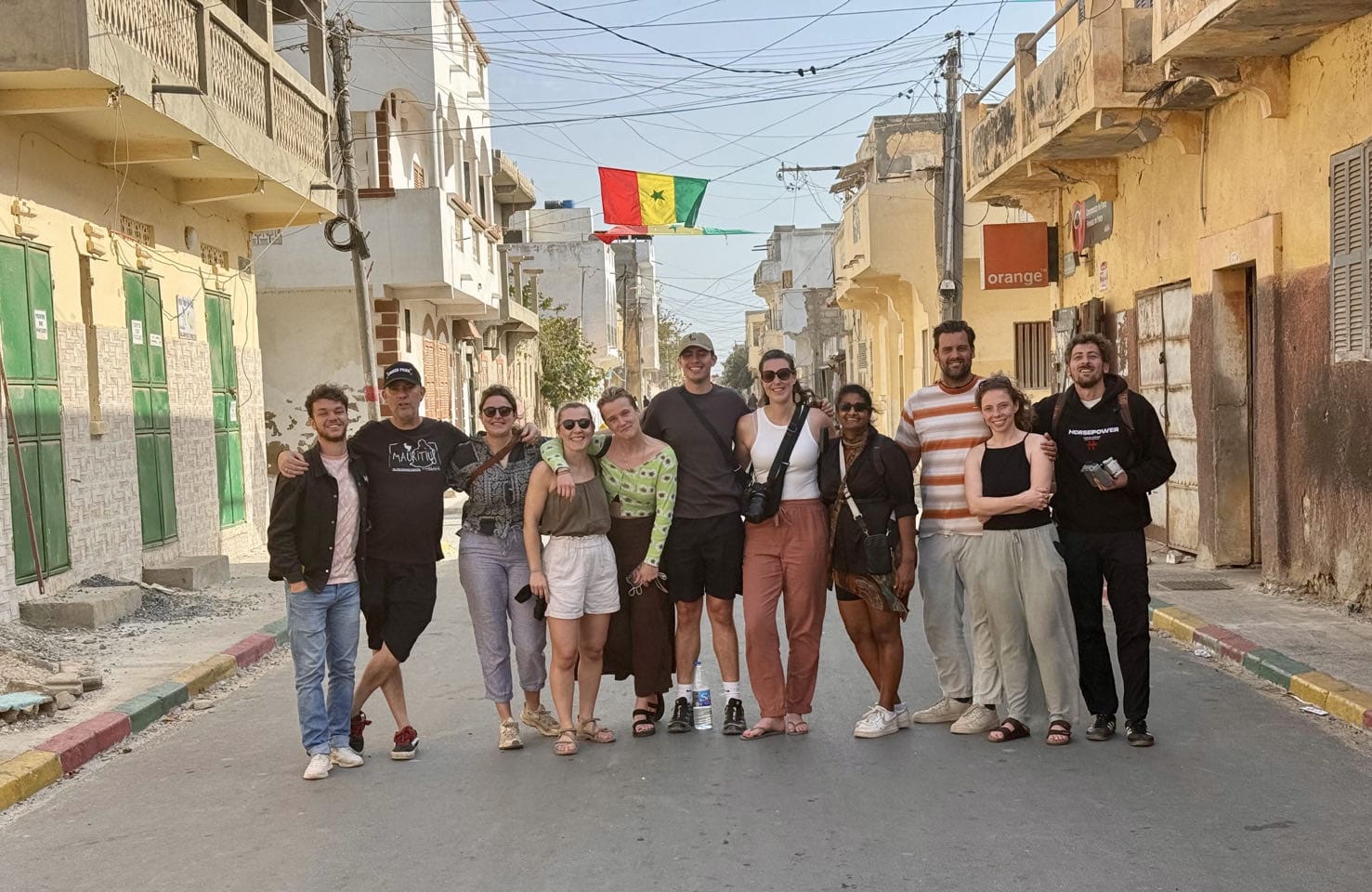 Een groep van twaalf mensen staat glimlachend midden op een zonovergoten straat, met de armen om elkaar heen. Achter hen hangt een Senegalese vlag boven de weg te midden van gele gebouwen.