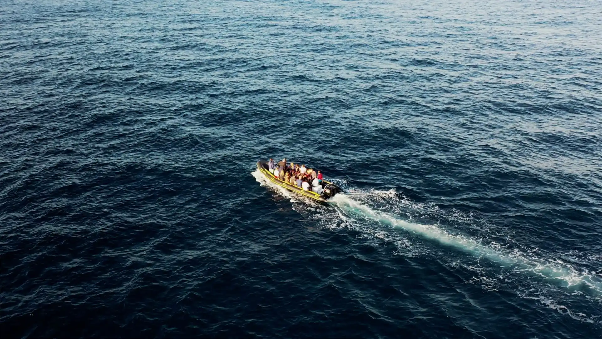 Een kleine overvolle boot met verschillende mensen aan boord vaart over diepblauw, open water en laat een wit kielzog achter. De uitgestrekte zee omringt de boot en benadrukt zijn isolement.