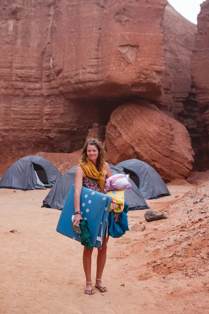 Een vrouw staat op zandgrond voor grijze tenten en grote rode rotsformaties, draagt een blauw slaapmatje en kleurrijk beddengoed, draagt een sjaal en sandalen. Het lijkt een camping in een canyon in de woestijn.