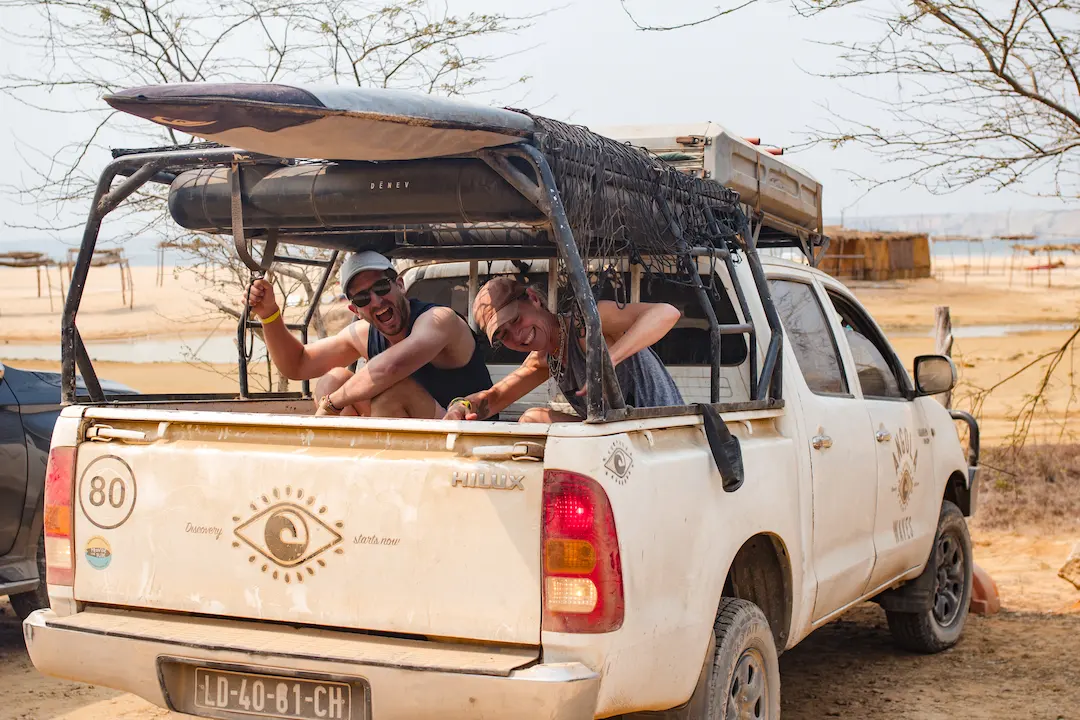 Twee lachende mannen zitten achterin een stoffige witte pick-up met surfplanken op het dak, geparkeerd op een zandstrand. Op de achtergrond staan droge bomen, strandhutjes en een wazige lucht.