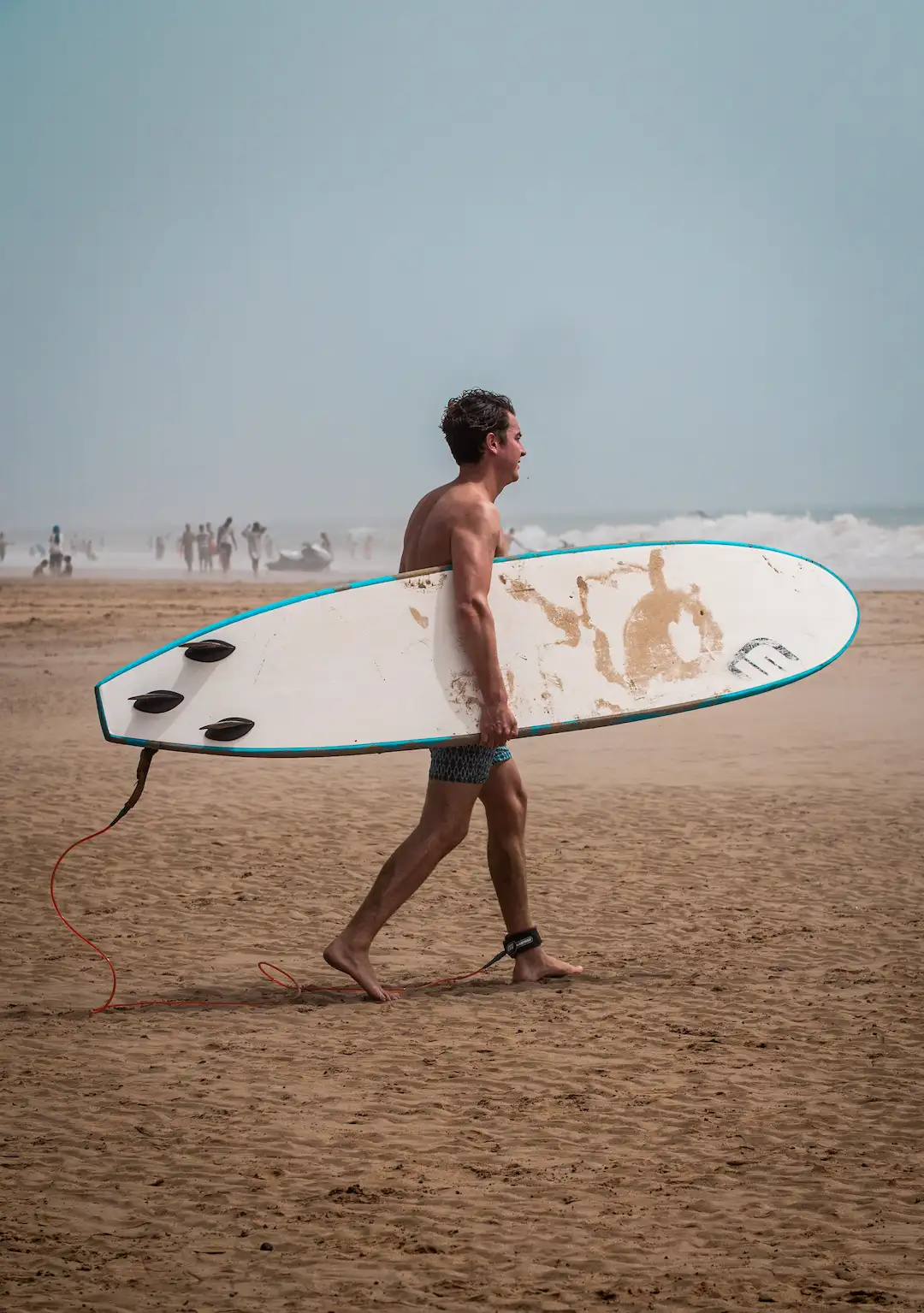 Een man in zwembroek loopt op blote voeten over een zandstrand met een surfplank onder zijn arm. Op de surfplank ligt wat zand. Op de achtergrond zijn mensen en golven te zien.