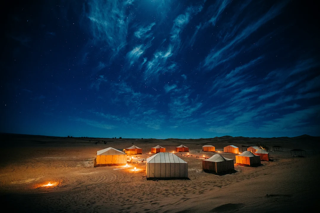 Een groep verlichte tenten en kleine kampvuurtjes staat 's nachts opgesteld in een zanderige woestijn, onder een hemel vol sterren en pluizige wolken, met daarboven de maan die helder schijnt.