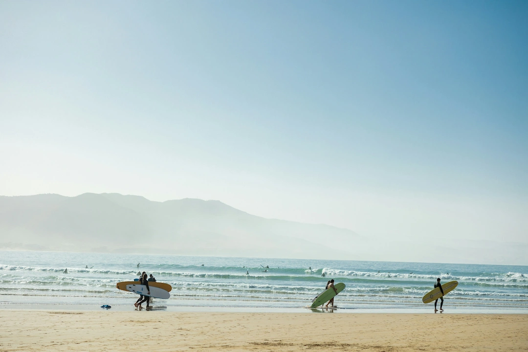 Verschillende mensen lopen met surfplanken over een zandstrand richting de zee; in het water zijn golven en surfers te zien en op de achtergrond zijn bergen te zien onder een helderblauwe hemel.