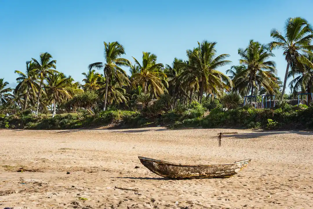 Een verweerde houten boot ligt op een zandstrand, met op de achtergrond palmbomen en groen, onder een helderblauwe hemel.