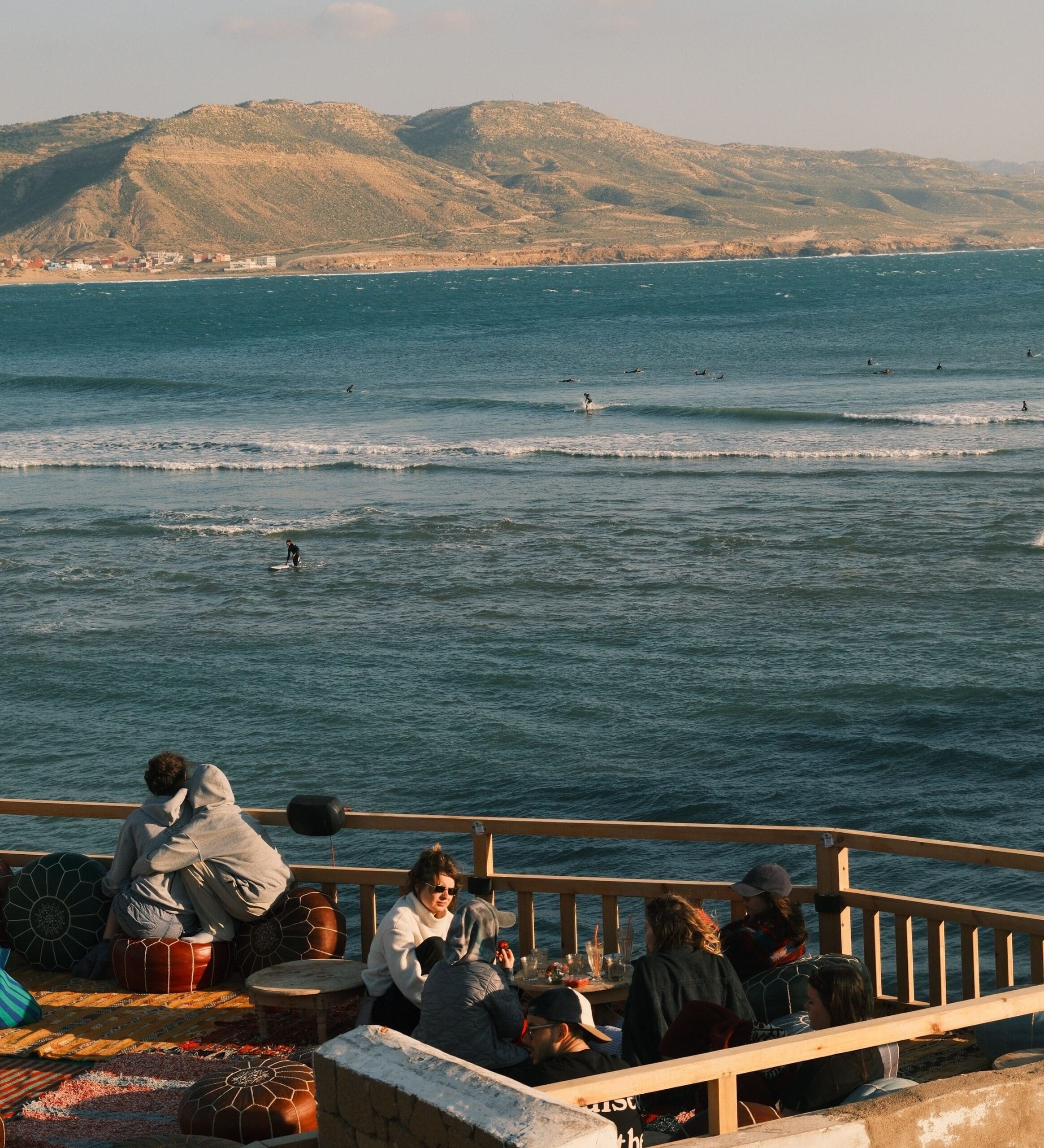 Mensen zitten op een terras met uitzicht op de oceaan, met surfers in het water en bergen op de achtergrond. De groep lijkt te ontspannen en gezellig te kletsen in het warme zonlicht.