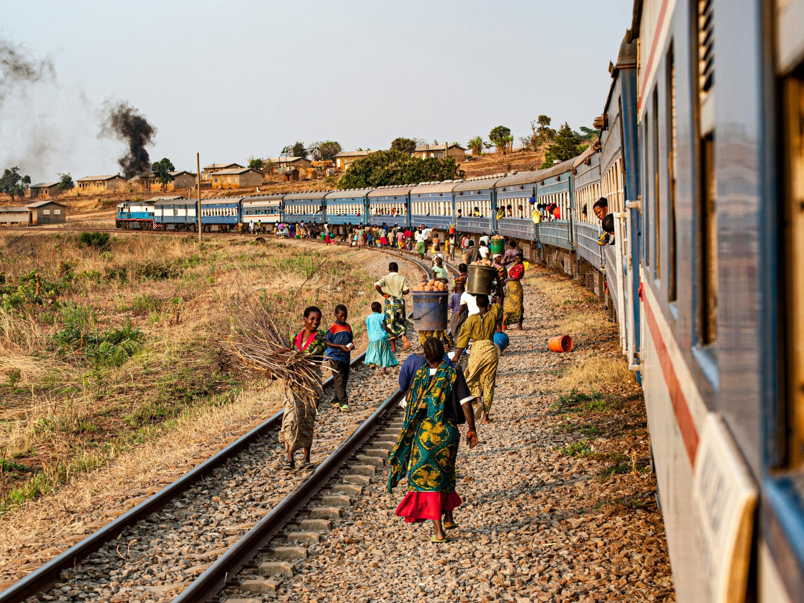 Mensen lopen langs een gebogen spoorweg naast een lange blauw-witte trein; sommigen dragen goederen of bundels takken. Onder een nevelige hemel zijn een landelijk landschap en een dorp in de verte te zien.