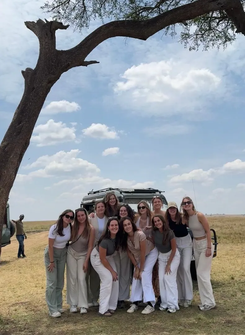 Een groep van twaalf vrouwen poseert en lacht onder een boom in een grasveld, met op de achtergrond een safarivoertuig; de lucht is gedeeltelijk bewolkt.