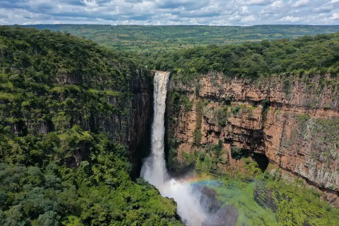 Een hoge waterval stort zich neer langs een rotswand, omgeven door een weelderig groen bos; aan de voet ervan zijn mist en een vage regenboog te zien onder een gedeeltelijk bewolkte hemel.