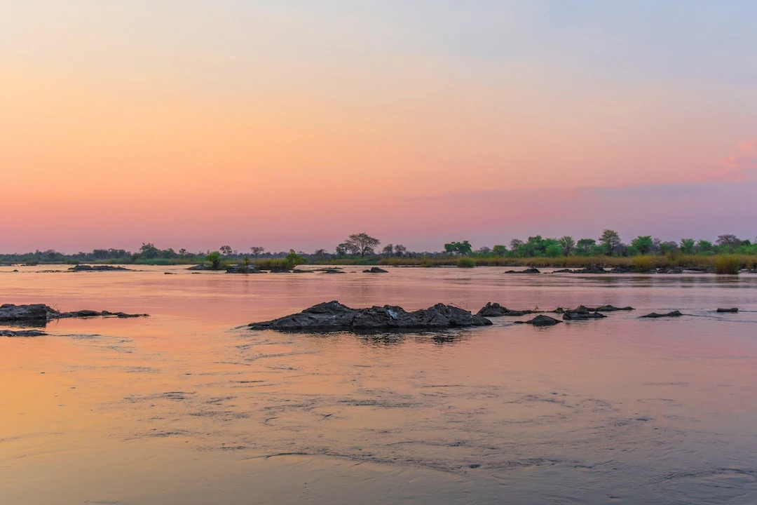 Een brede rivier bij zonsondergang, met kalm water waarin roze en oranje tinten weerspiegelen, her en der verspreide rotsen en groene begroeiing langs de oever in de verte, onder een pastelkleurige hemel.