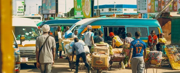 Op een drukke openluchtmarkt duwen mensen karren vol goederen tussen geparkeerde bussen en bestelwagens door, met reclameborden en winkels op de achtergrond.