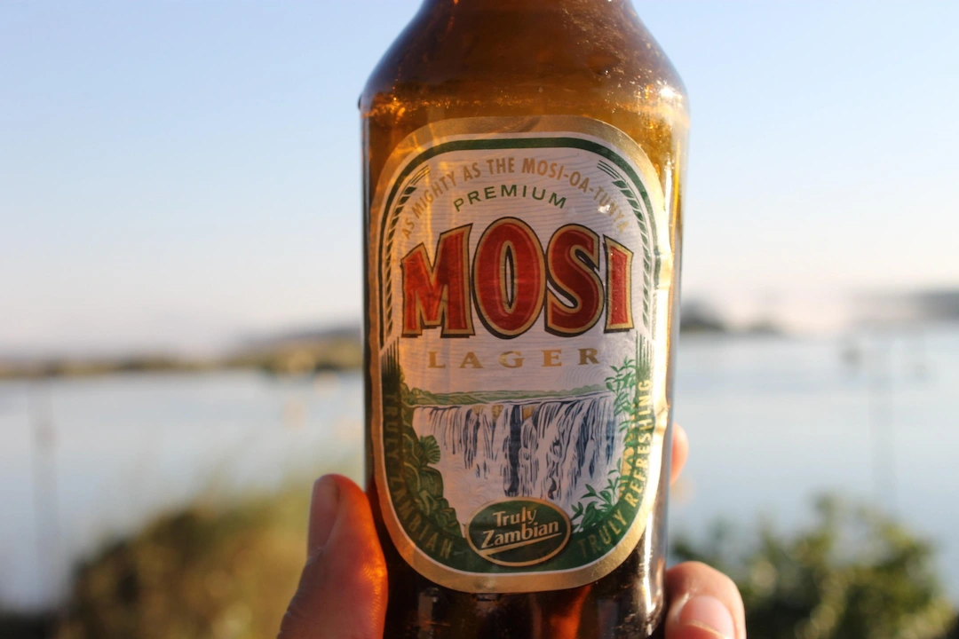 Een hand houdt een fles Mosi Lager-bier vast met een etiket waarop de Victoriawatervallen te zien zijn. De achtergrond is wazig en toont water en groen onder een stralende hemel.