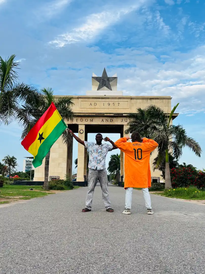 Twee mensen poseren voor de Onafhankelijkheidsboog van Ghana. De ene houdt een Ghanese vlag vast, terwijl de andere een oranje shirt draagt met het nummer 10 op de rug. Op de achtergrond zijn palmbomen en een blauwe lucht te zien.