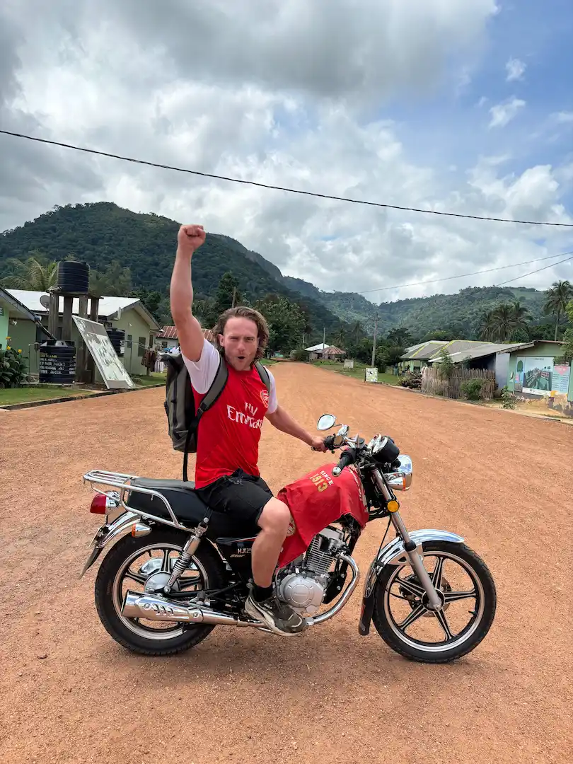 Een persoon in een rood Arsenal-shirt en met een rugzak zit op een motorfiets op een onverharde weg en steekt zijn vuist in de lucht, met op de achtergrond groene heuvels, huizen en een bewolkte hemel.
