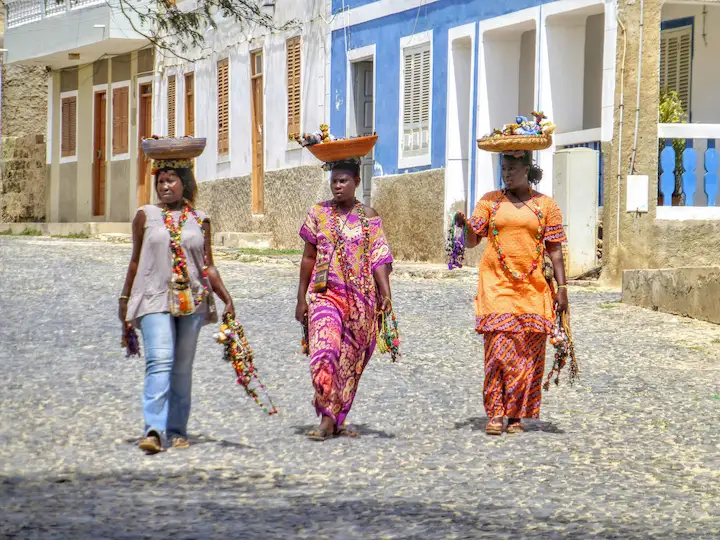 Drie vrouwen lopen over een straat met kasseien, met manden op hun hoofd en kleurrijke voorwerpen in hun handen, met witte en blauwe gebouwen op de achtergrond.