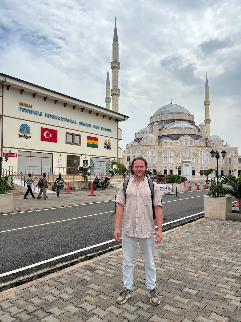 Een man die op het trottoir staat voor het gebouw van de Hudai Tiwnikli International Senior High School en een grote moskee met twee minaretten.