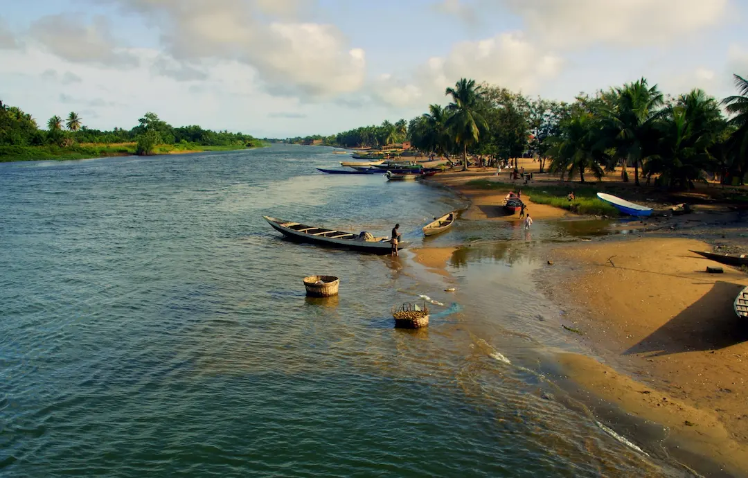 Een man staat in een houten boot aan de oever van een rivier die omzoomd is met palmbomen en waar verschillende kleine vissersboten liggen.