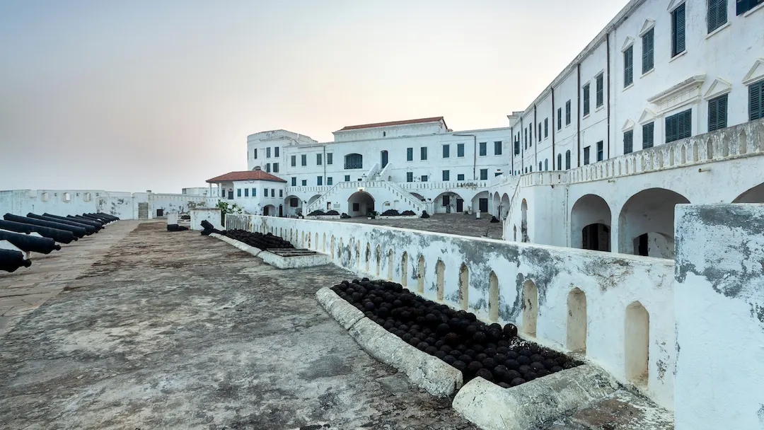 Een binnenplaats bij Cape Coast Castle, met witte gebouwen met booggewelven en rijen kanonnen en kanonskogels op een stenen wal.