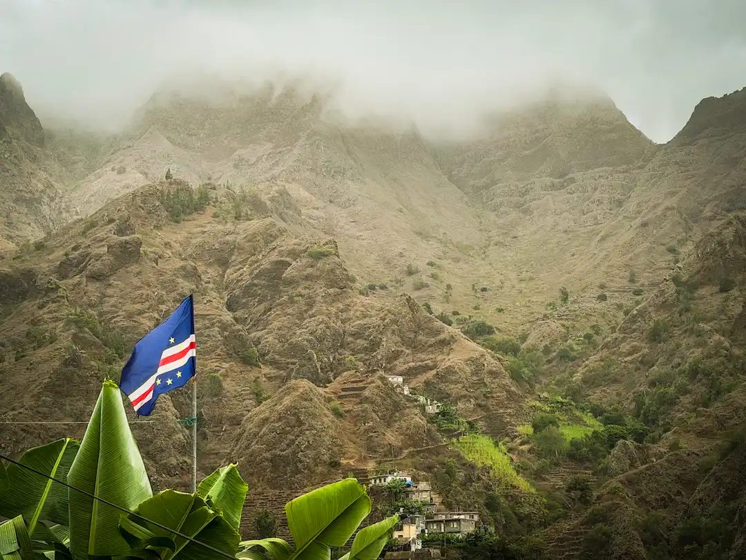 De vlag van Kaapverdië wappert tegen een achtergrond van torenhoge, in mist gehulde bergen en huizen op terrassen tegen de helling.