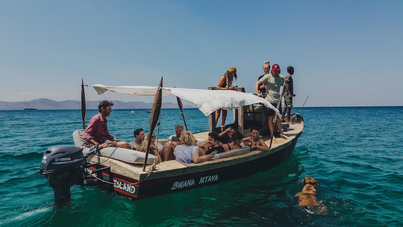 Een groep mensen zit op een kleine houten boot met de naam Bwana Mtaya midden in het turquoise water, terwijl er vlakbij een hond zwemt.