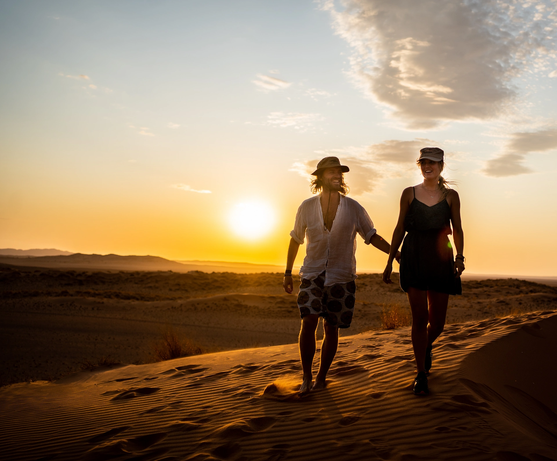 Een man en een vrouw met hoeden op, die bij zonsondergang hand in hand over een zandduin in de woestijn lopen.