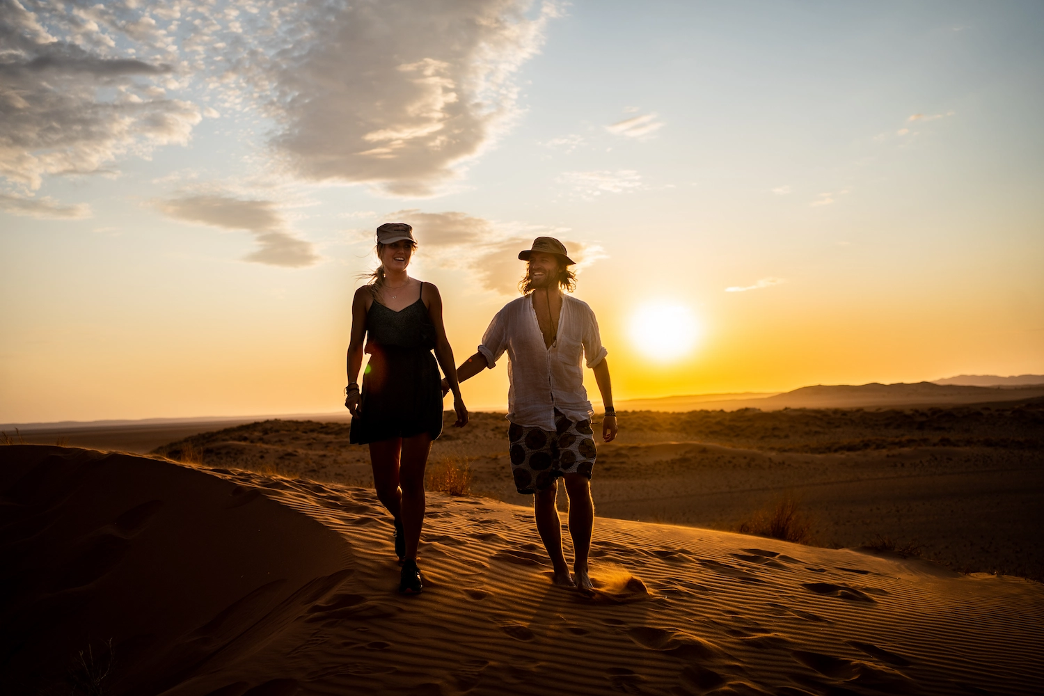 Een man en een vrouw lopen bij zonsondergang hand in hand over een zandduin in de woestijn.