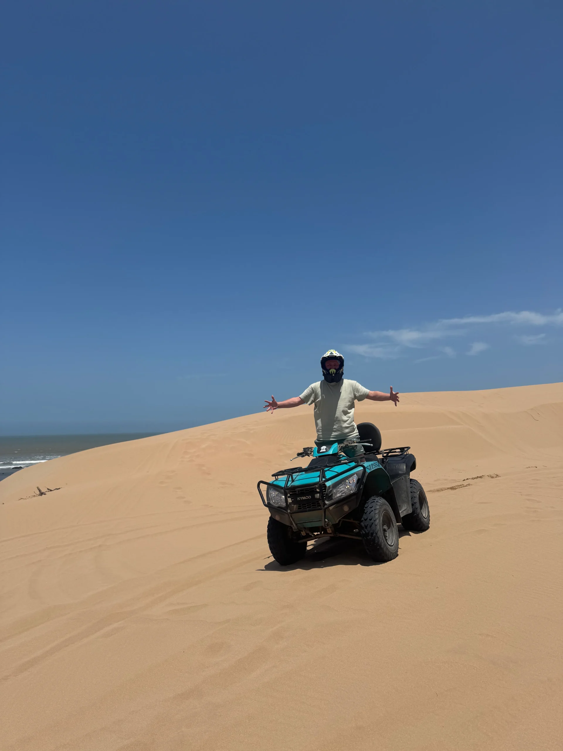 A person wearing a helmet and arms outstretched, riding a teal ATV on a sandy dune near the ocean.