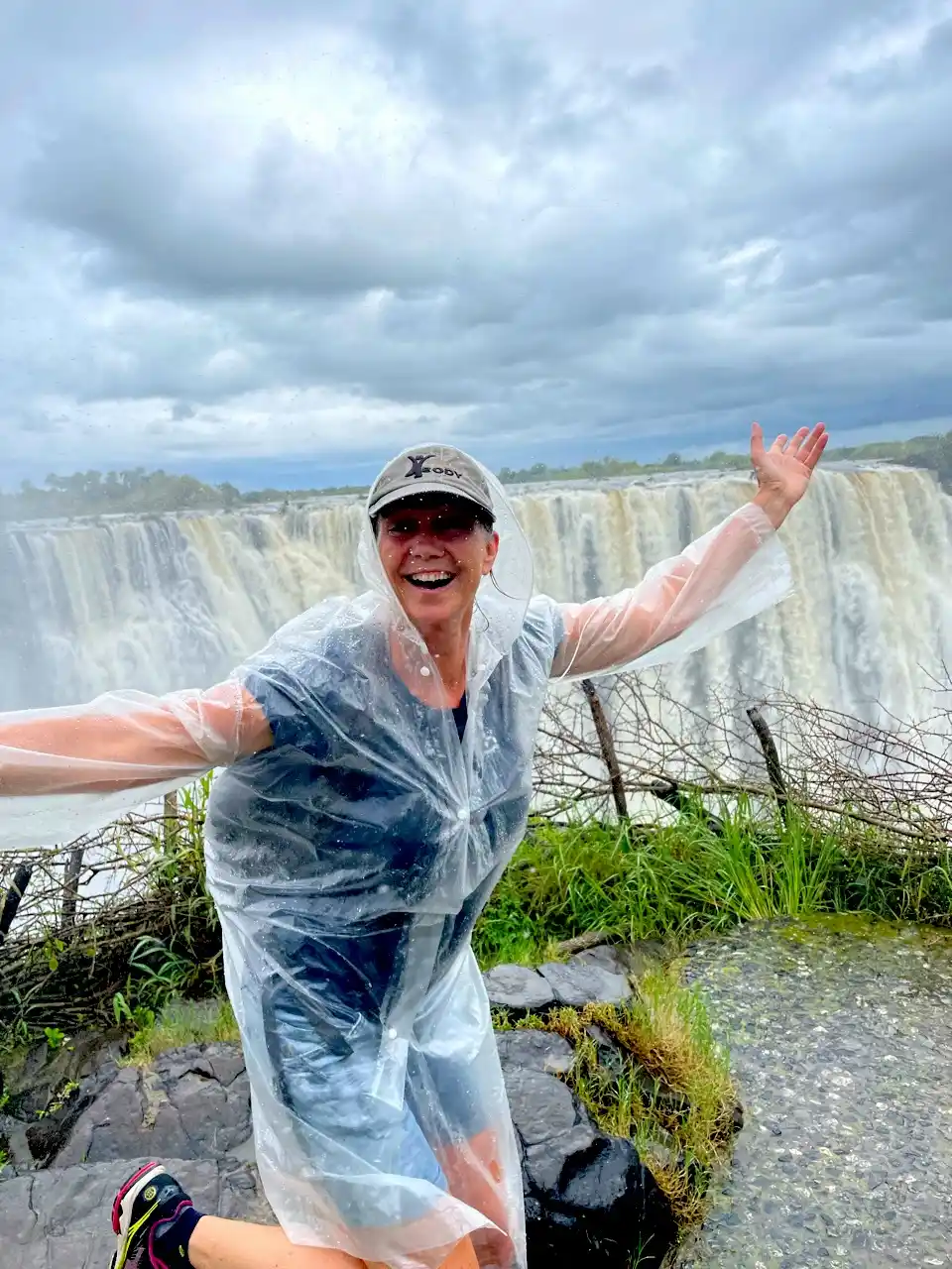 Een vrouw in een doorzichtige plastic regenjas juicht met uitgestrekte armen voor een grote waterval.