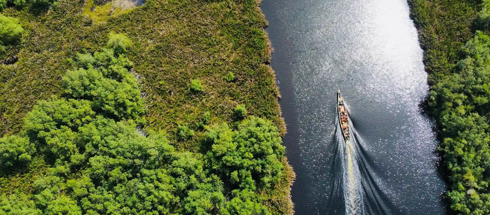 Een luchtfoto van een smalle boot die vaart over een rivier, omringd door weelderige groene begroeiing.