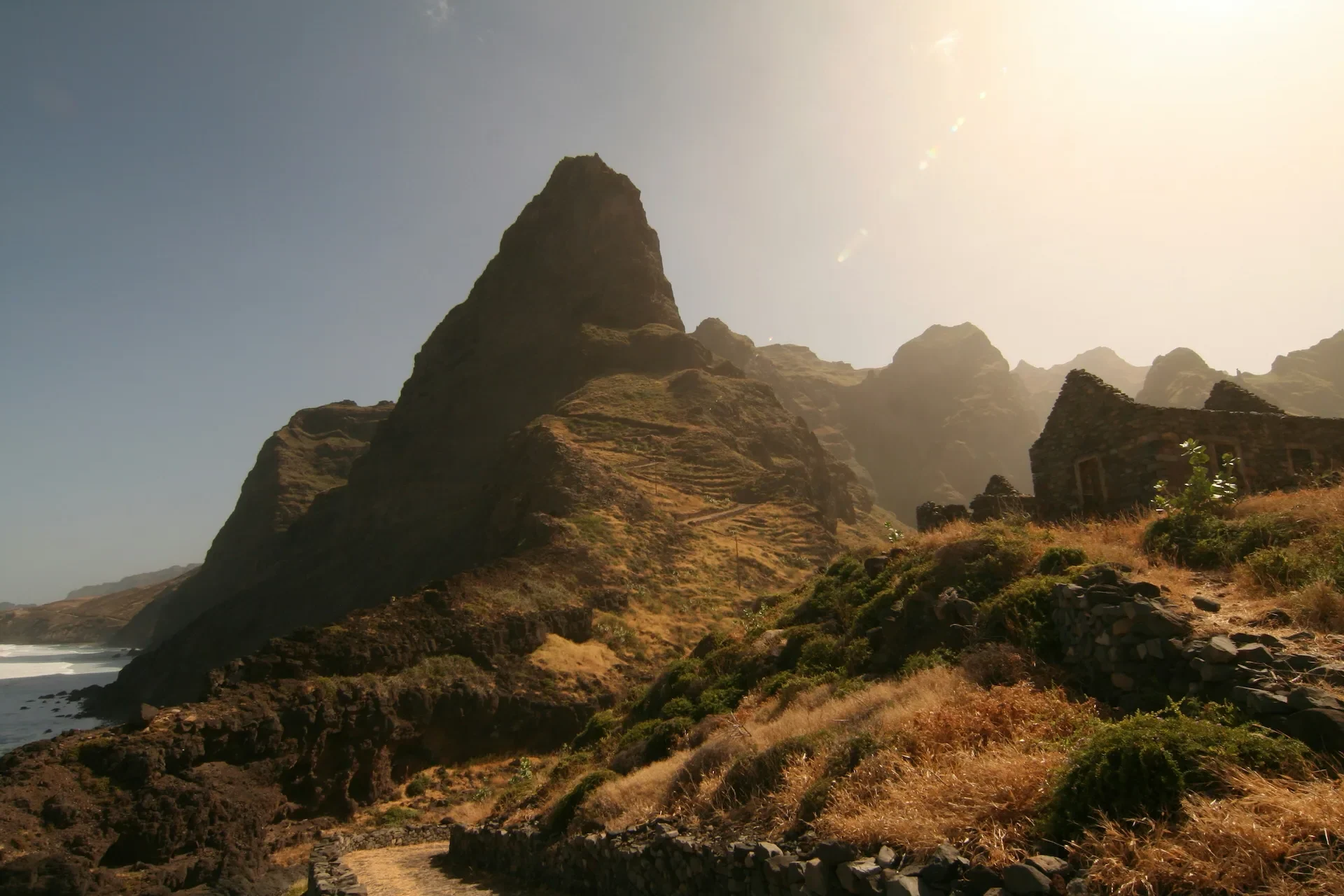 A rocky mountain peak towers over a dirt path and the ruins of a stone building near a rugged coastline.