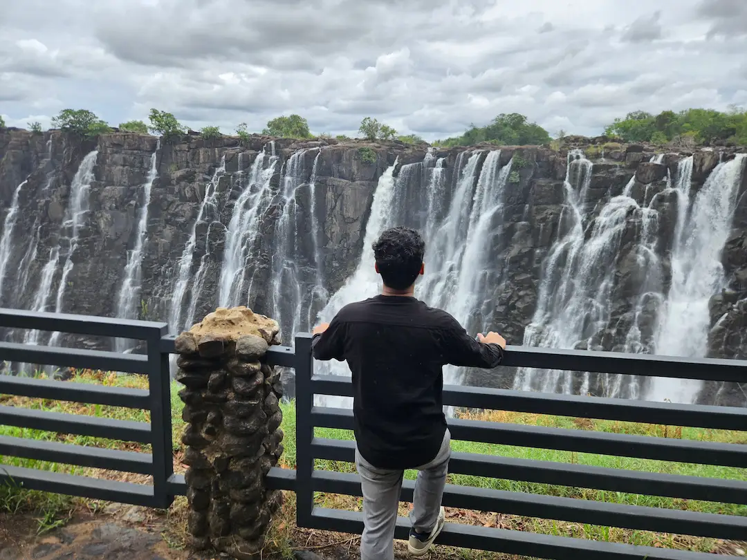 Een persoon, van achteren gezien, leunt tegen een zwarte reling en kijkt uit over een brede, uit meerdere niveaus bestaande waterval.